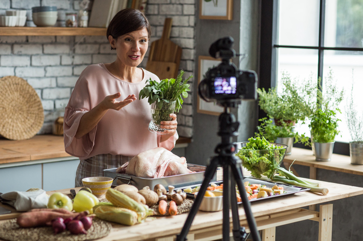 Une caméra sur un trépied filme une femme qui prépare de la nourriture dans une cuisine en regardant la caméra et en parlant tout en tenant à la main des herbes fraîches.