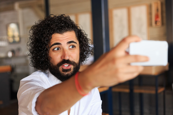 Un jeune homme aux cheveux foncés regarde attentivement un écran de téléphone cellulaire qu'il tient comme s'il voulait prendre une photo ou une vidéo de lui.