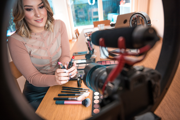 Une jeune femme bien habillée et s’étant bien maquillée est assise à une table où elle montre des produits cosmétiques et tient des bâtons de rouge à lèvres devant une caméra.